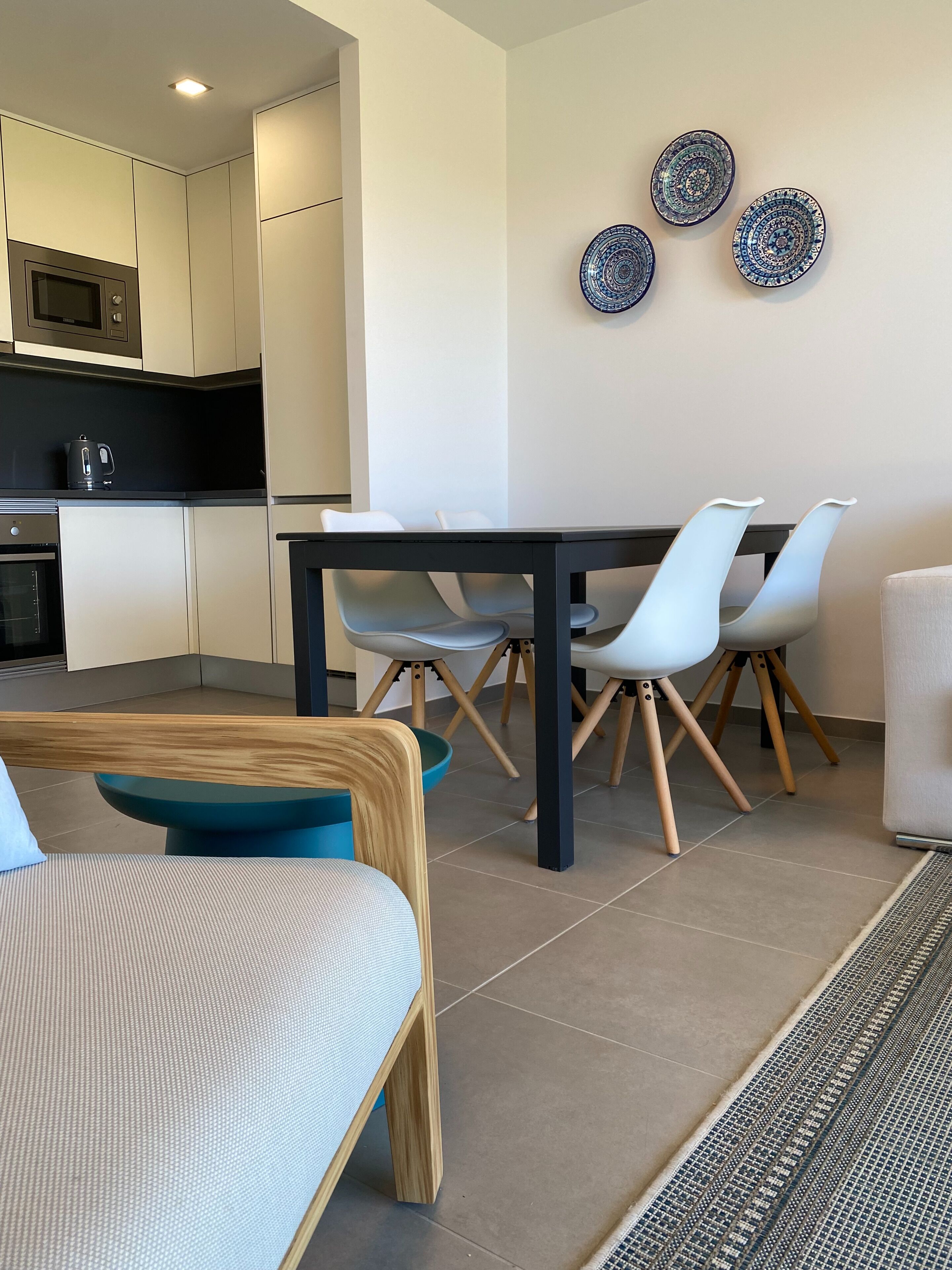 Una cocina moderna con armarios blancos, encimera negra y un hervidor. Hay una zona de comedor con una mesa negra, sillas blancas y un tazón azul.