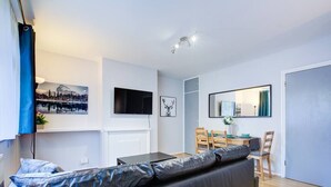 A modern living room with a black leather sofa, a flat-screen TV mounted on the wall, a dining area with a table and chairs, and a large mirror reflecting the room. - Statera Apartments - Studio Apartment in Walworth (London)