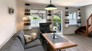 Un salon chaleureux avec un canapé, une table en bois et une chaise. Il y a une fenêtre avec des rideaux, une lampe et une vue sur l’extérieur.