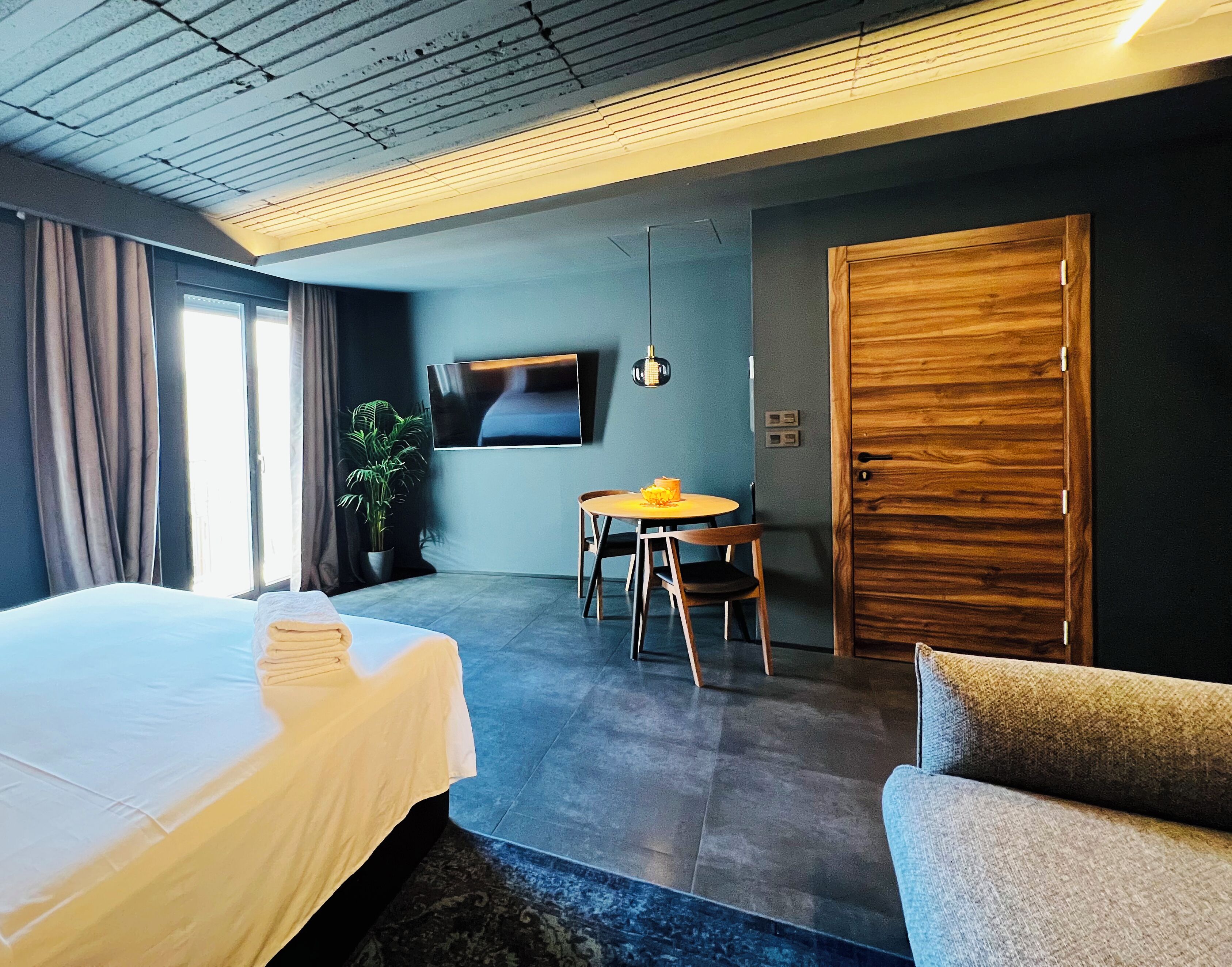 A modern hotel room with a large bed, a small round table with chairs, a wooden door, and a potted plant.