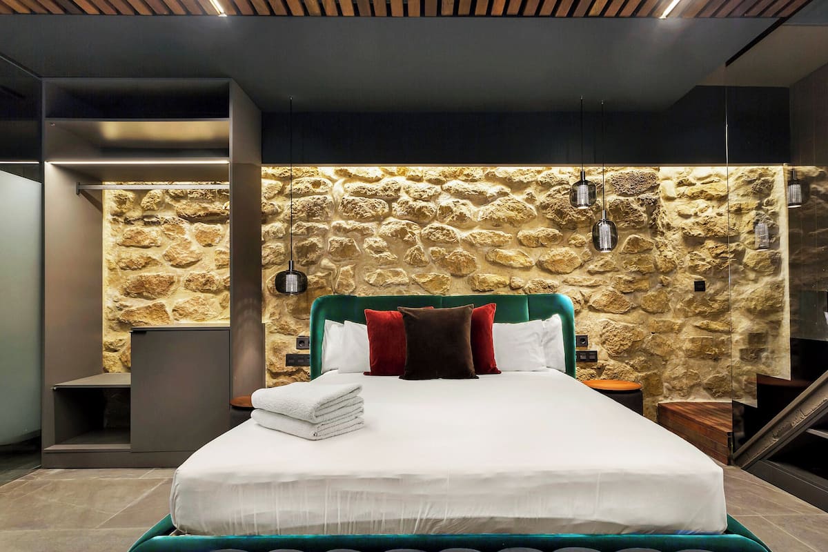 A modern bedroom with a stone wall, a green upholstered headboard, and a neatly made bed with white linens and multiple pillows.