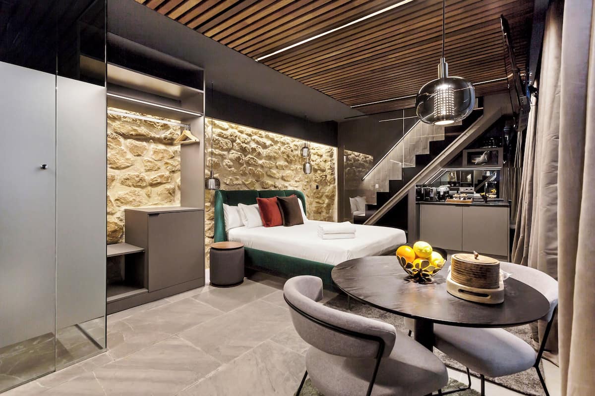 A modern hotel room with a stone accent wall, a bed, a dining area with a table and chairs, and a staircase leading to an upper level.