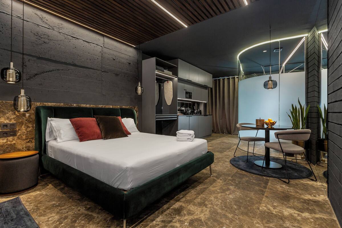 A modern bedroom with a large bed, a desk with chairs, and a built-in kitchen area.