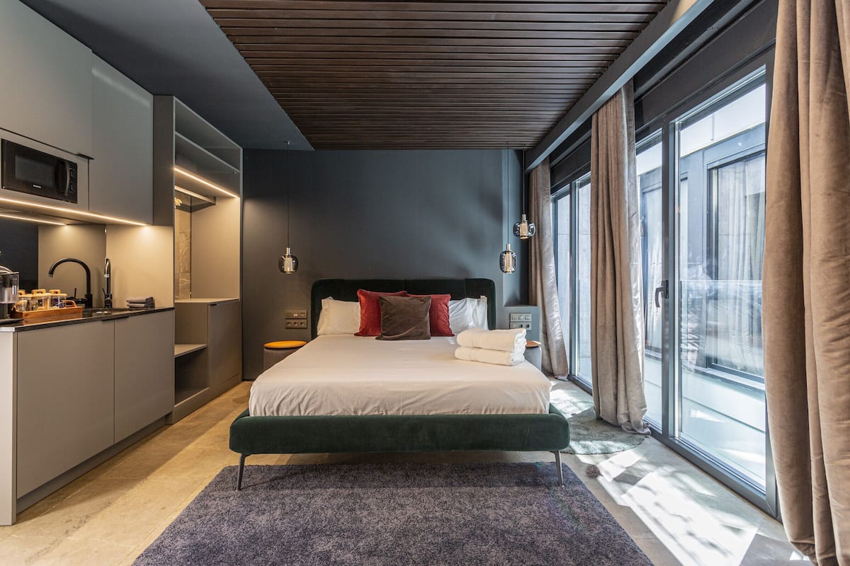 A modern bedroom with a large bed, a built-in wardrobe, a kitchenette, and a large window with curtains.