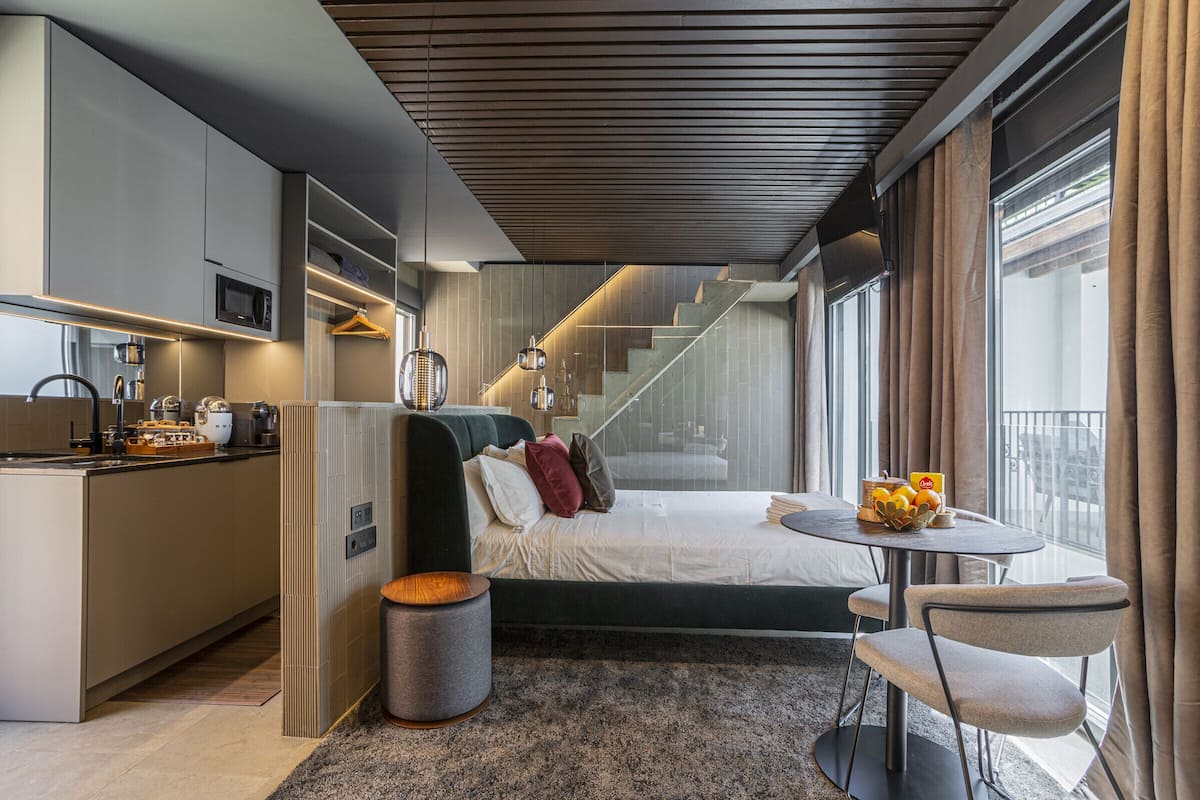 A modern hotel room with a bed, a dining table, a kitchen area, and a staircase.