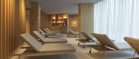 Un espacio de spa con sillones reclinables, una camilla de masaje y una pared con estantes que contienen libros y objetos decorativos.