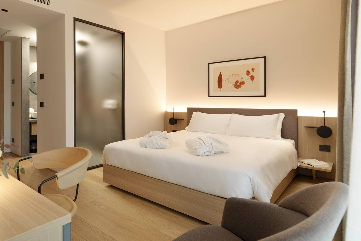 A modern hotel room with a neatly made bed, a desk with a chair, and a bedside table with a lamp.