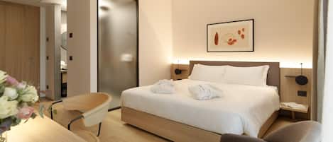 A modern hotel room with a neatly made bed, a desk with a chair, and a bedside table with a lamp.