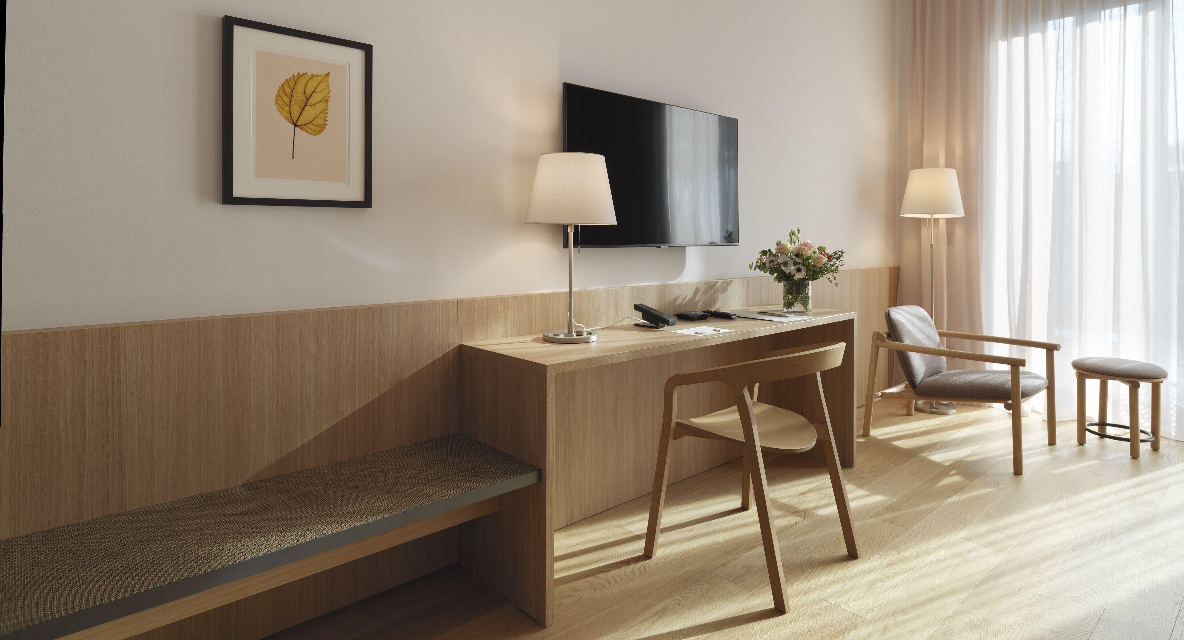 A modern hotel room with a wooden desk, a flat-screen TV, a bench, and two chairs.