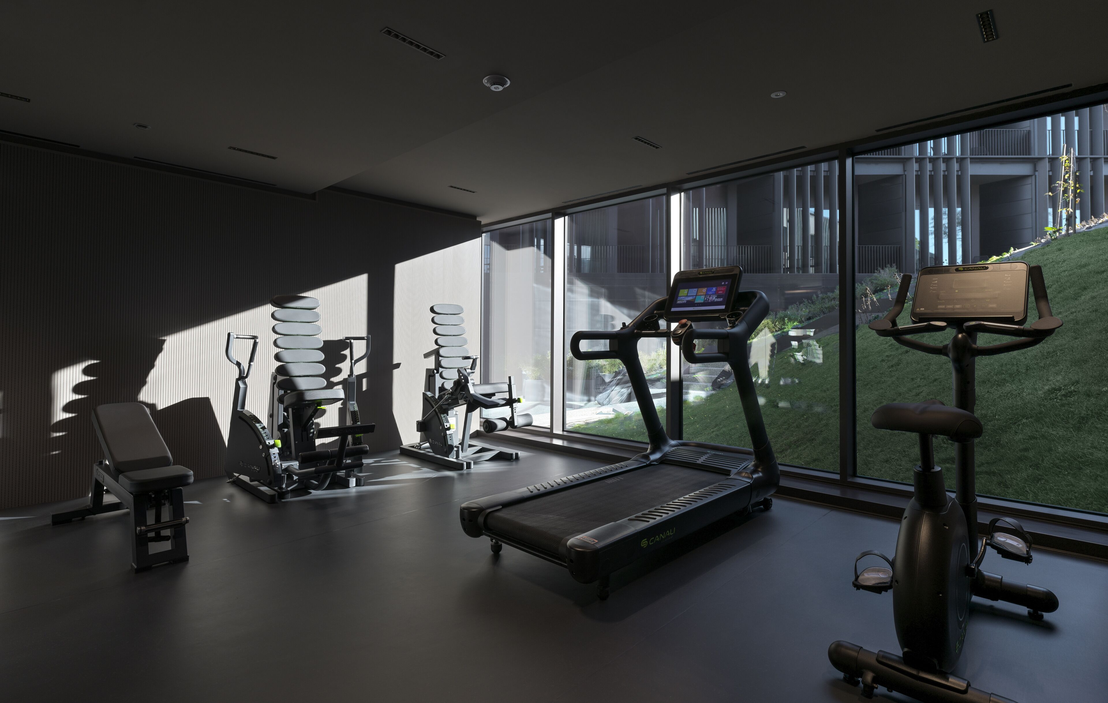 Un gimnasio moderno con diversos equipos de ejercicio, ventanas amplias y vistas a un edificio cercano.
