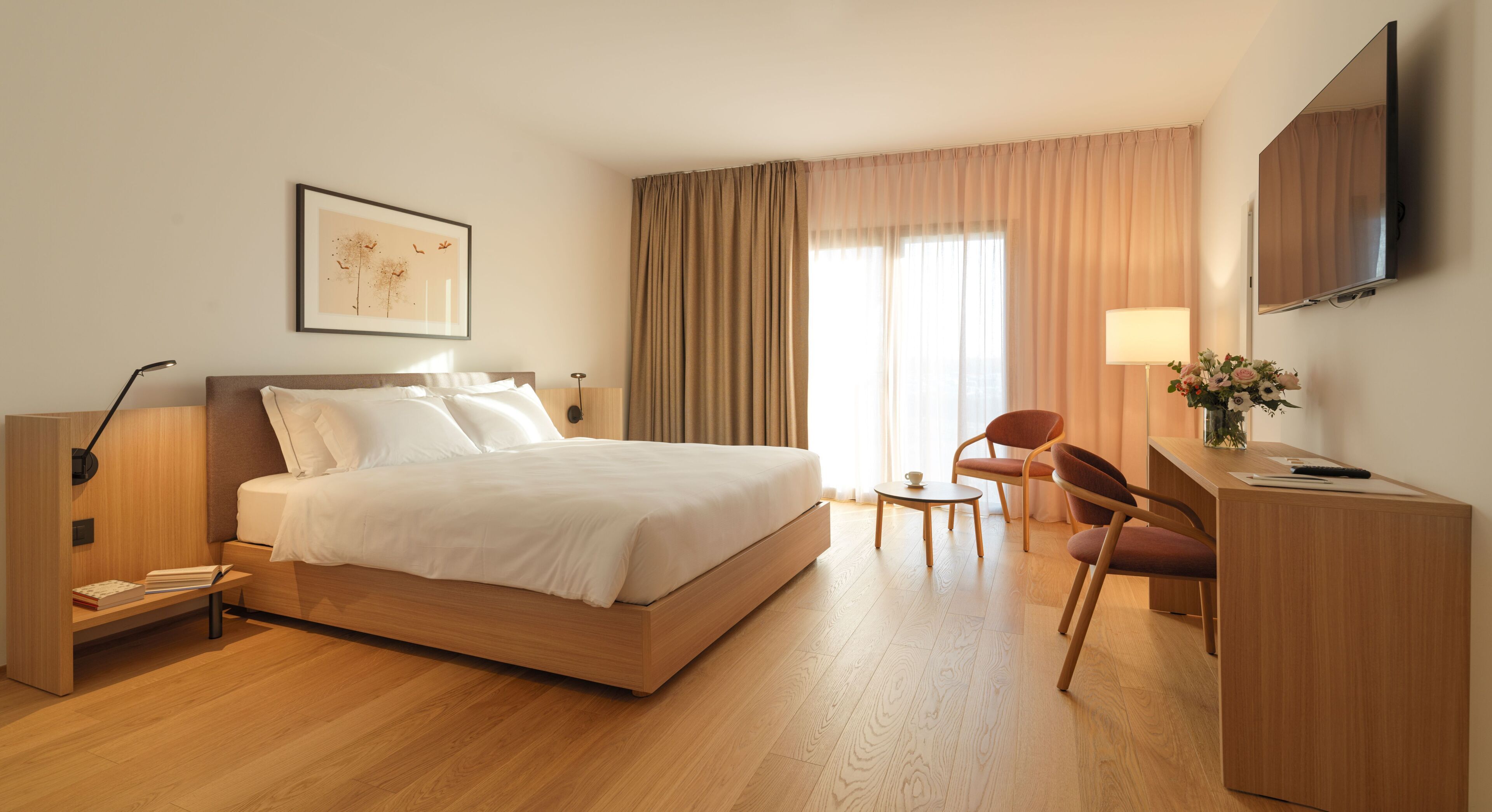 A hotel room with a large bed, a desk, a chair, a lamp, and a TV mounted on the wall.