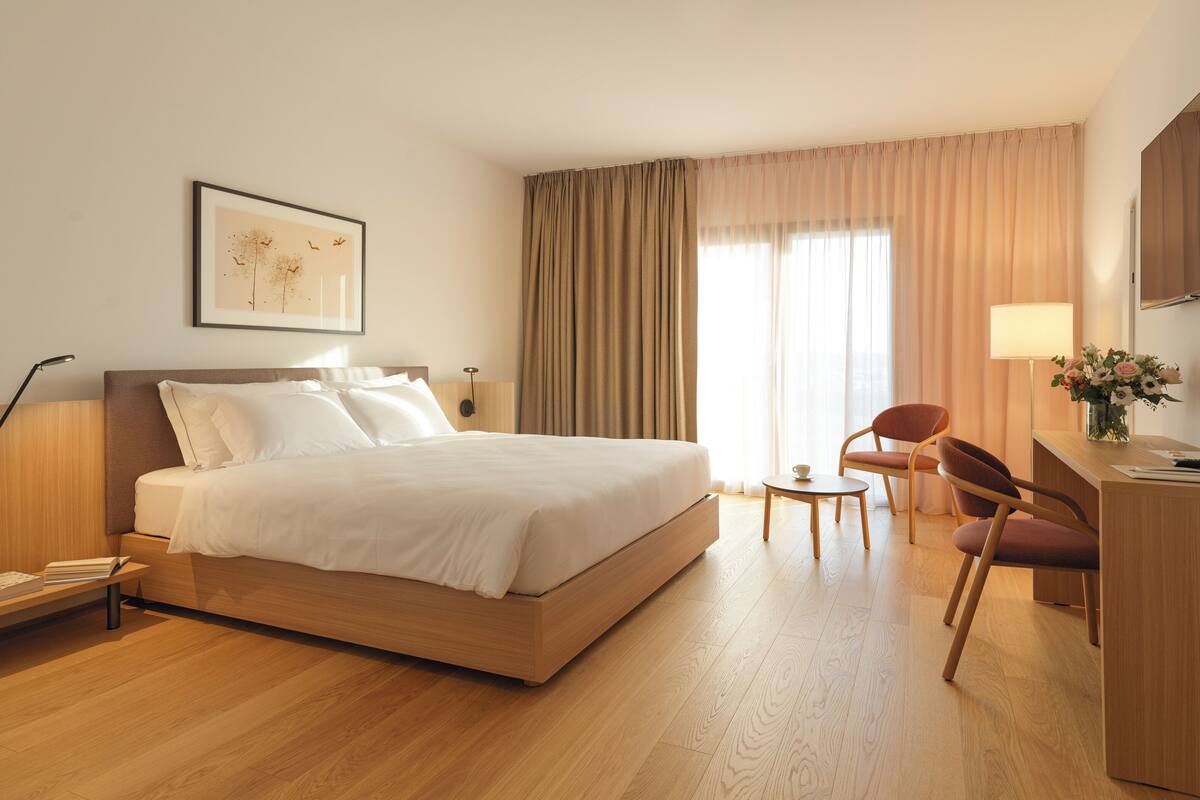 A hotel room with a large bed, a desk with a lamp, a chair, and a television on the wall.