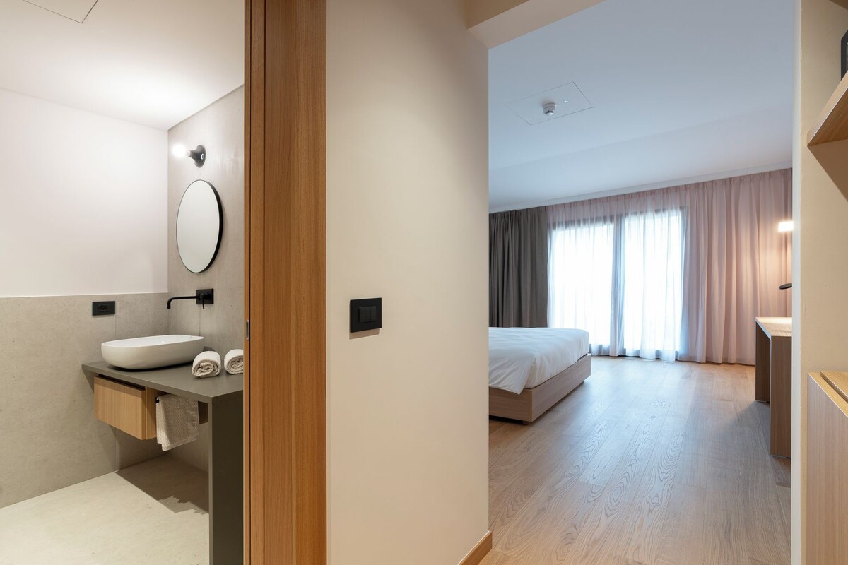 Une chambre d’hôtel moderne avec un plancher en bois, un grand lit, une salle de bain dotée d’un lavabo et d’un miroir, et une vue sur l’extérieur grâce à de grandes fenêtres.