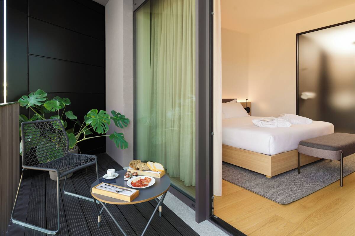 A modern hotel room with a bed, bedside table, and a small balcony area with a chair and potted plants.