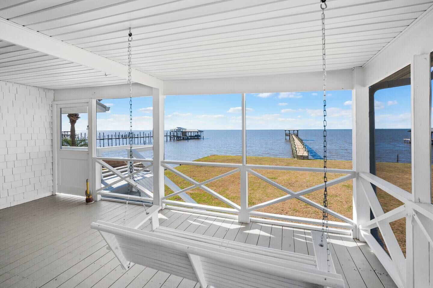 Une terrasse en bois blanche avec une balançoire, une vue sur la mer et une jetée en arrière-plan.