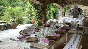 Una larga mesa de comedor preparada para una comida en un patio al aire libre con muros de piedra y vegetación.