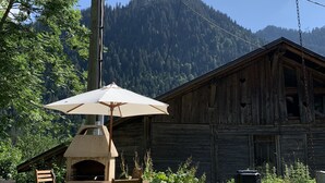 Un entorno rústico al aire libre con una mesa y sillas de madera bajo un gran paraguas, un horno de leña y una gallina en primer plano.