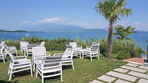 Een terras met witte loungestoelen en een palmboom, met uitzicht op een rustige zee met boten en verre bergen.