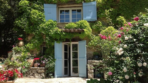 Ein rustikales Steinhaus mit Holztür und blauen Fensterläden, umgeben von üppiger Vegetation und blühenden Pflanzen.