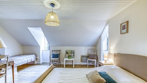 Et rummeligt soveværelse på loftet med en stor seng, to mindre senge, en stol, et bord og en hængende lampe.