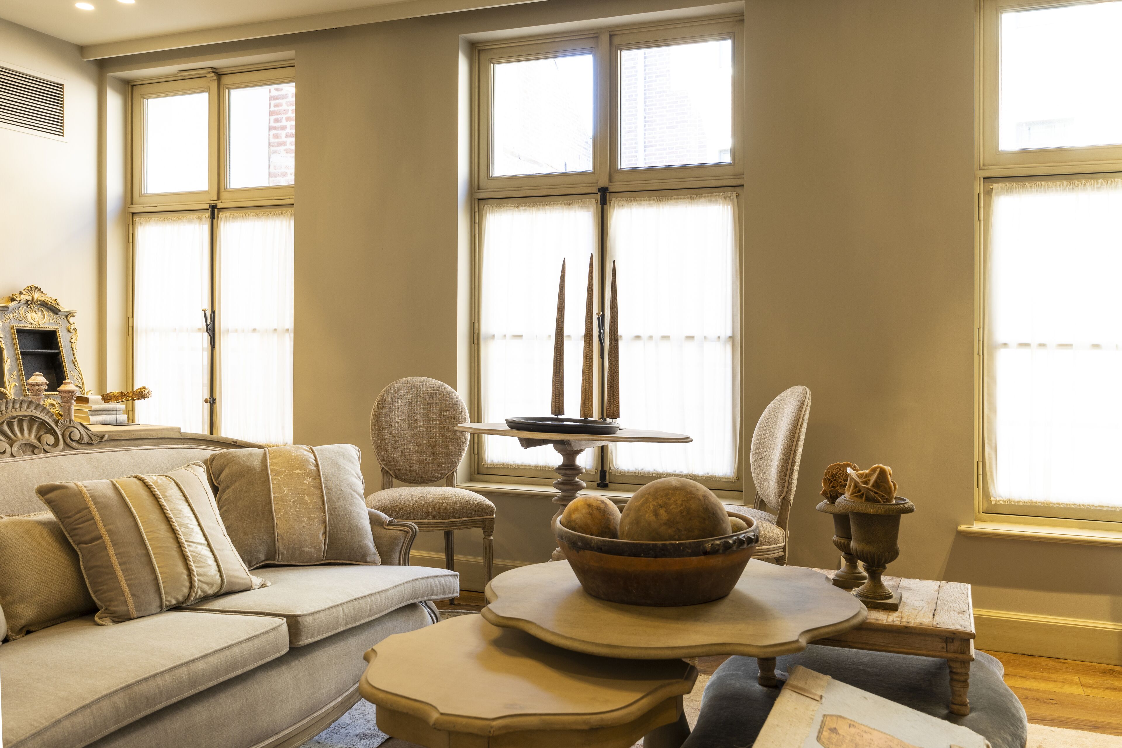 A living room with a beige sofa, two armchairs, a coffee table with decorative items, and a window with curtains.