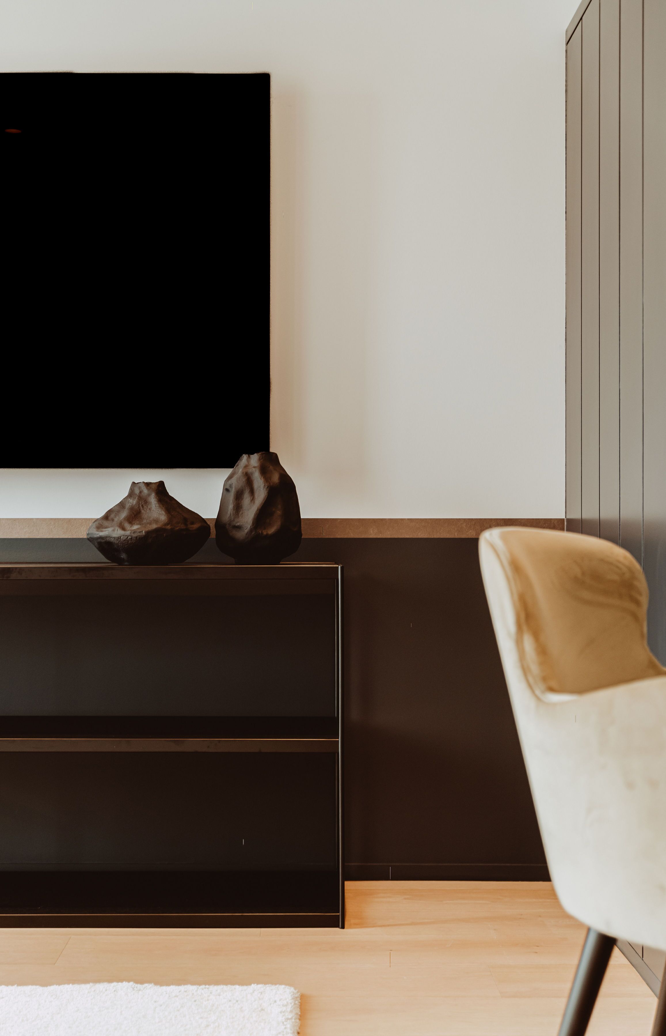 A modern living room with a flat-screen TV mounted on the wall, a dark wooden console table with two decorative items, a white cushioned chair, and a light-colored rug on the wooden floor.