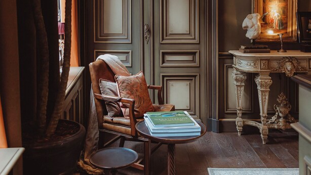 Una habitación con una planta grande en maceta, un sillón de cuero, una pequeña mesa con libros, una lámpara decorativa y un cuadro en la pared.