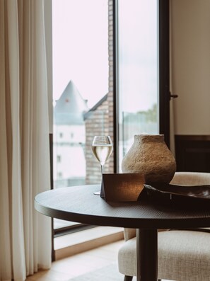 A table with a glass of white wine, a rustic vase, and a wooden tray in a room with large windows and a view of buildings. - Cobergher Hotel (Kortrijk)