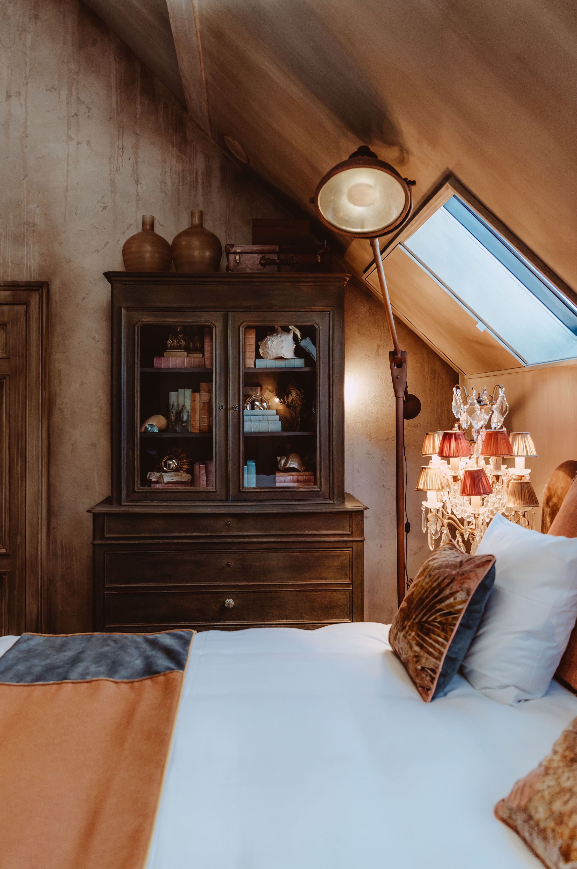 A bedroom with a skylight, a cabinet with glass doors, a chandelier, and a bed with pillows.