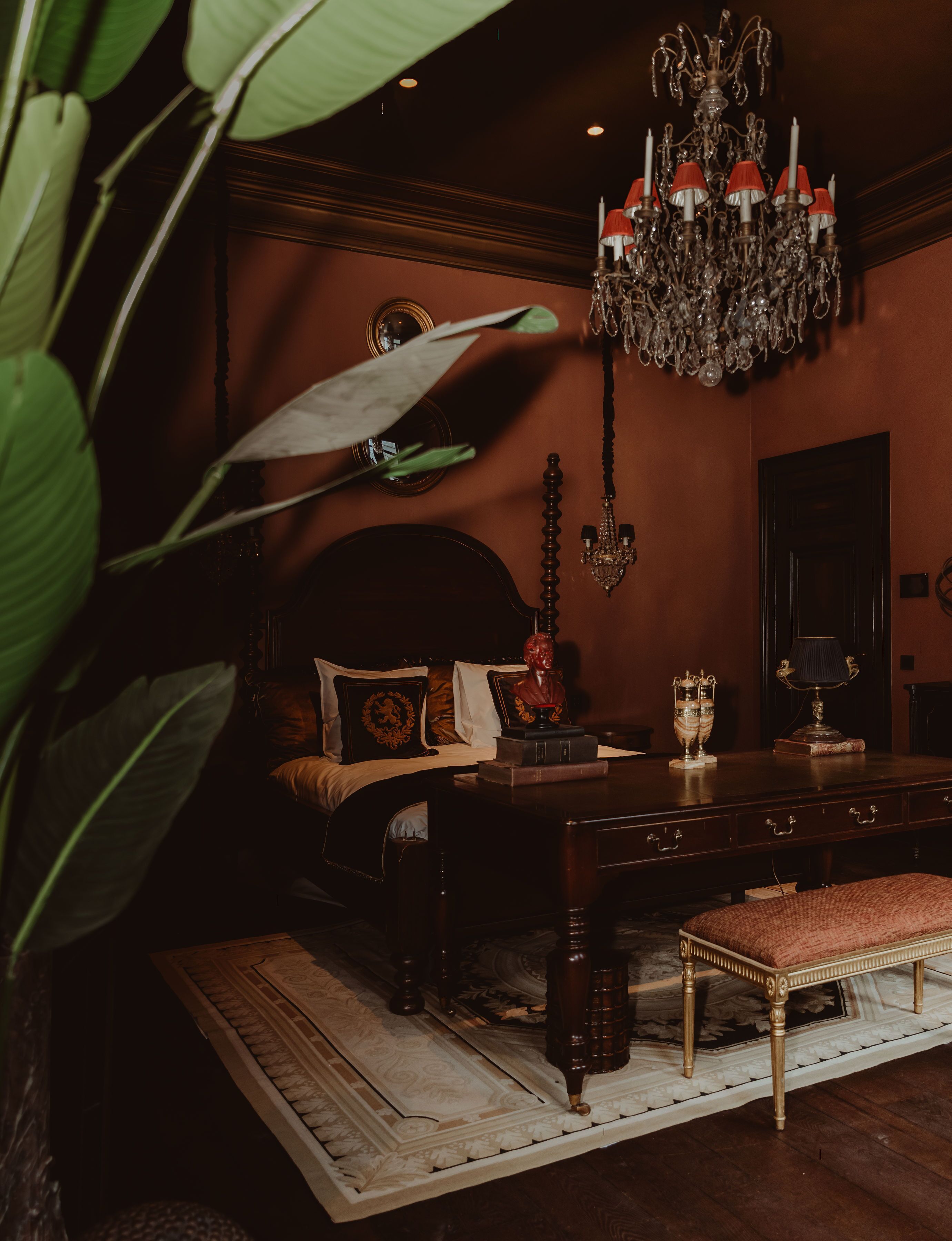 A dimly lit room with a large chandelier, a bed with ornate pillows, a wooden desk, and a large plant in the foreground.