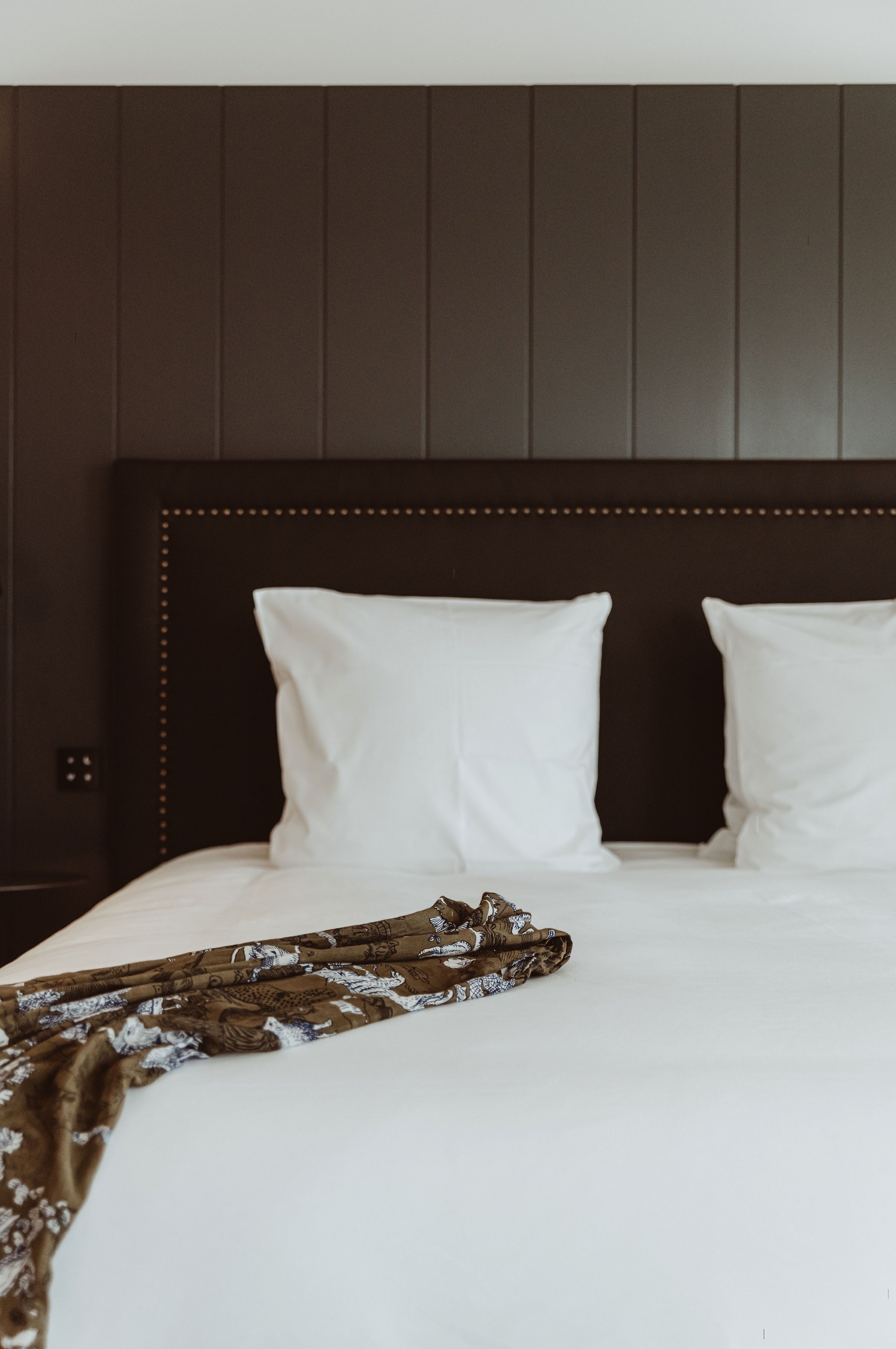 Een net opgemaakt bed met witte lakens en een bedektje met een patroon, voor een donker houten hoofdbord met decoratieve rand.