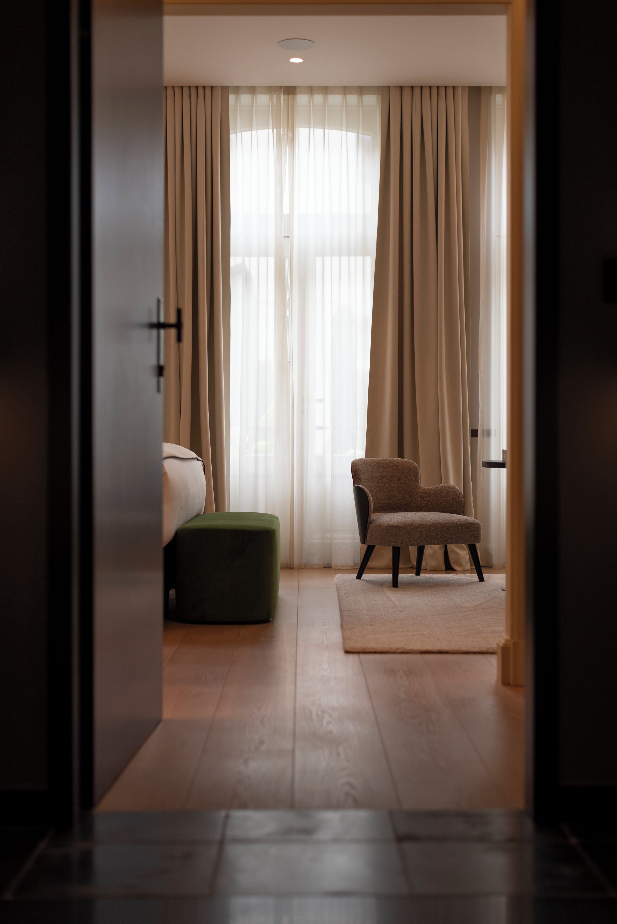 A hotel room with a bed, a green ottoman, a chair, and a view through sheer curtains.