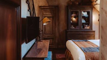 Een gezellige kamer met een houten plafond, een bed met een beddekleed met patroon, een houten kast met glazen deuren, en een tafel met een lamp.