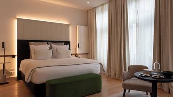 Habitación de hotel moderna con una cama grande, un taburete verde, una mesa redonda con una cúpula de vidrio y vistas de la ciudad a través de cortinas translúcidas.