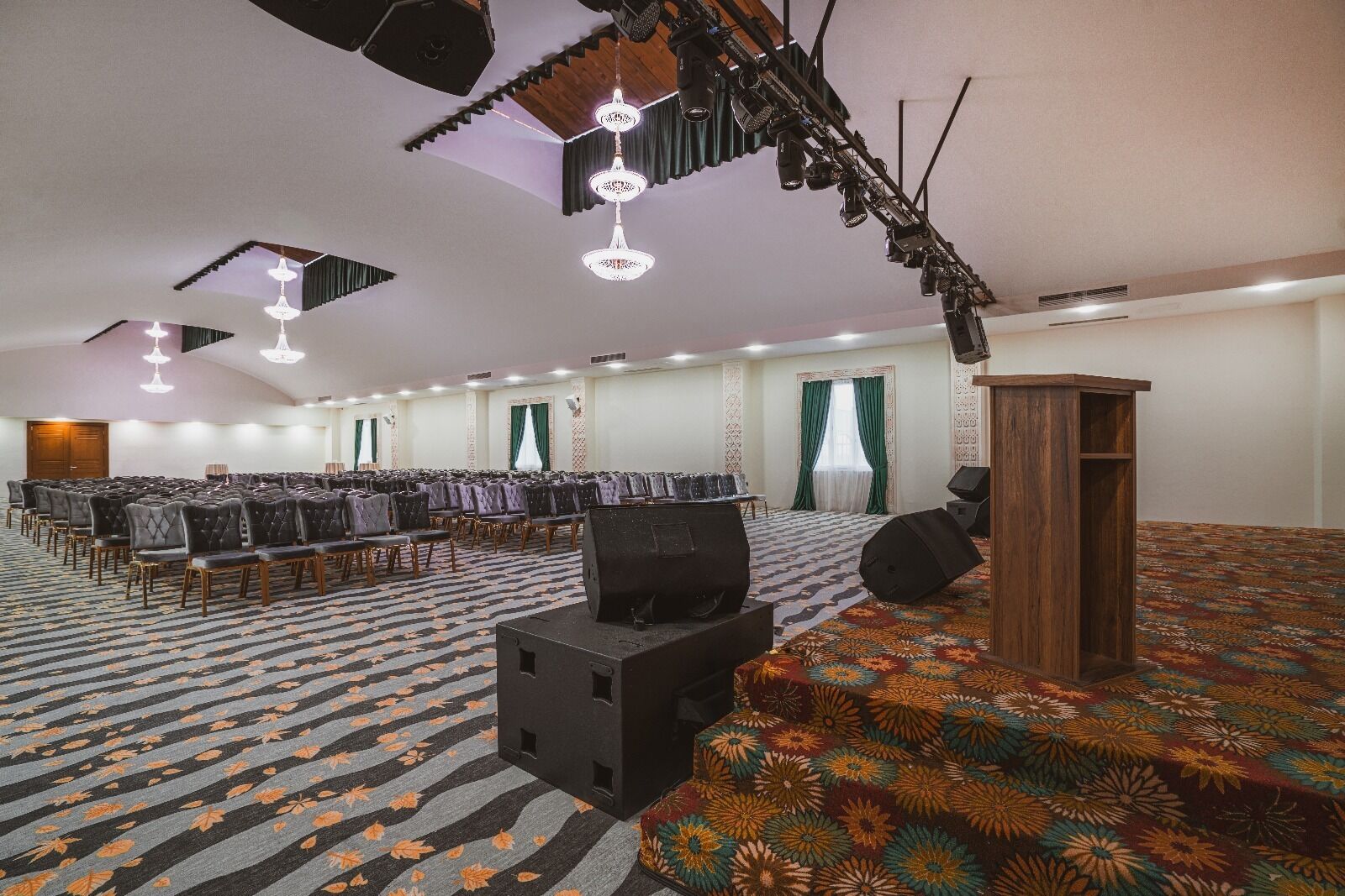 A conference room with rows of chairs, a stage with speakers, and a patterned carpet.