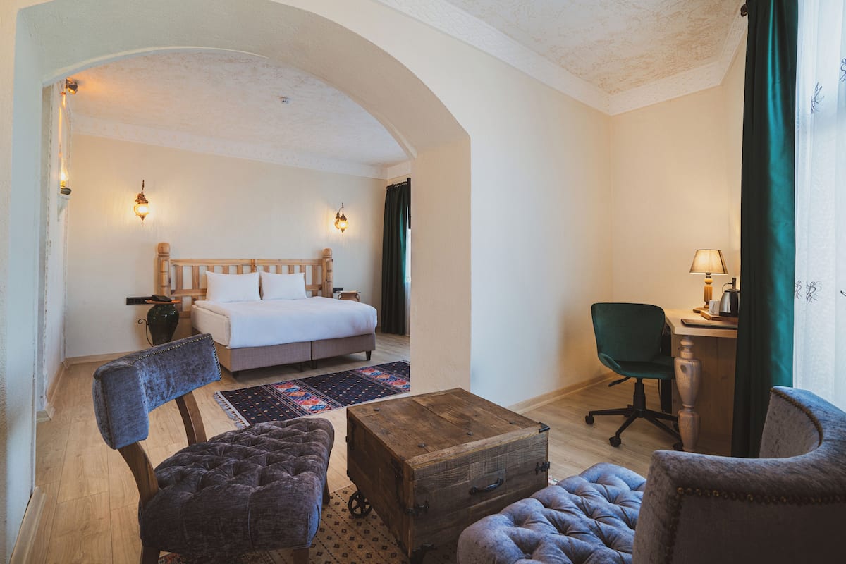 A hotel room with a bed, two armchairs, a wooden chest, a desk with a lamp, and a view through an arched doorway.