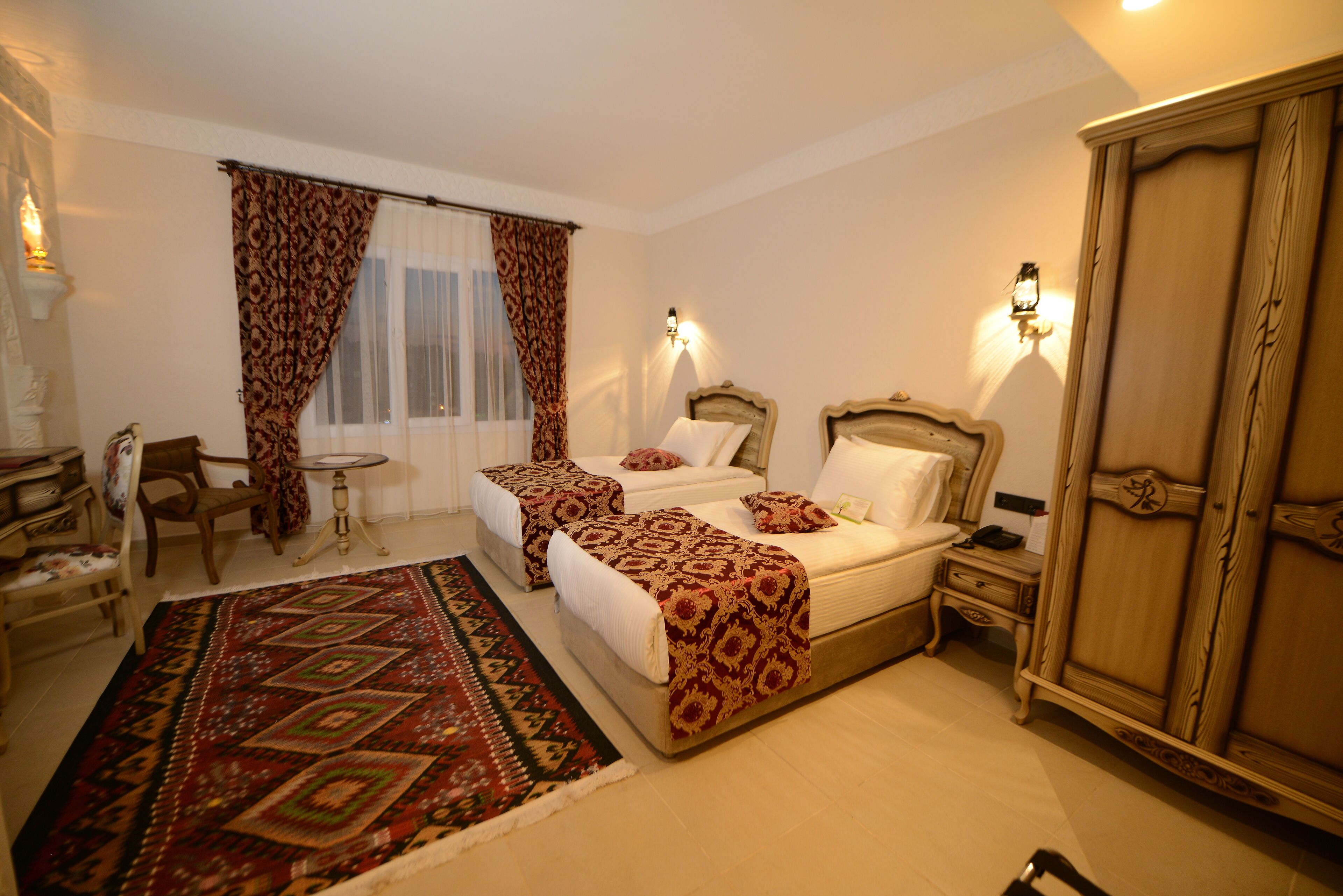 Habitación de hotel con dos camas, un armario de madera grande, una mesita pequeña y una alfombra estampada.
