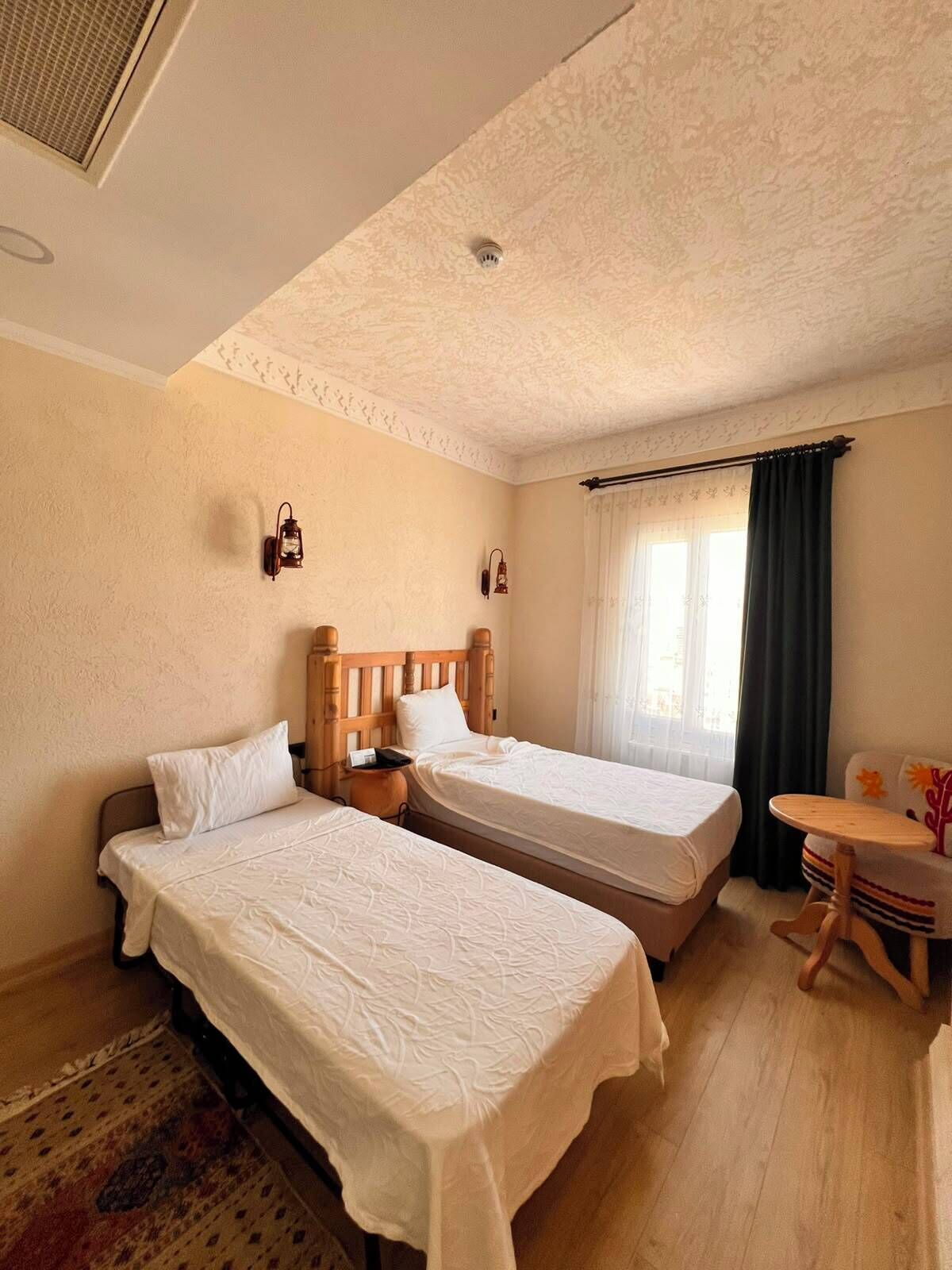A room with two beds, a small table, a lamp, and a window with curtains.