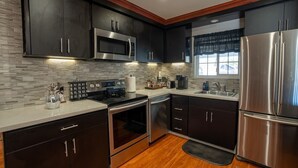 A modern kitchen with dark cabinetry, stainless steel appliances, a stone backsplash, and a wooden floor.