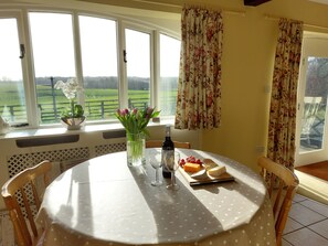 A dining area with a round table, chairs, a vase with flowers, a bottle, and a plate with cheese and fruit. - Meadow cottage- family friendly, two bedrooms both ensuite and all on one level. (Rushyford)