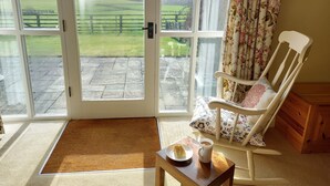 A room with a view of a green field through a glass door, a wooden chair with floral cushions, a small table with a plate and mug, and a wooden cabinet. - Meadow cottage- two bedrooms both ensuite and all on one level. (Rushyford)