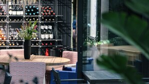 An outdoor seating area with a round wooden table and blue and red upholstered chairs. Behind the seating, there is a wine rack with various bottles. - Chapter 02 Gallery Suites (Heraklion)