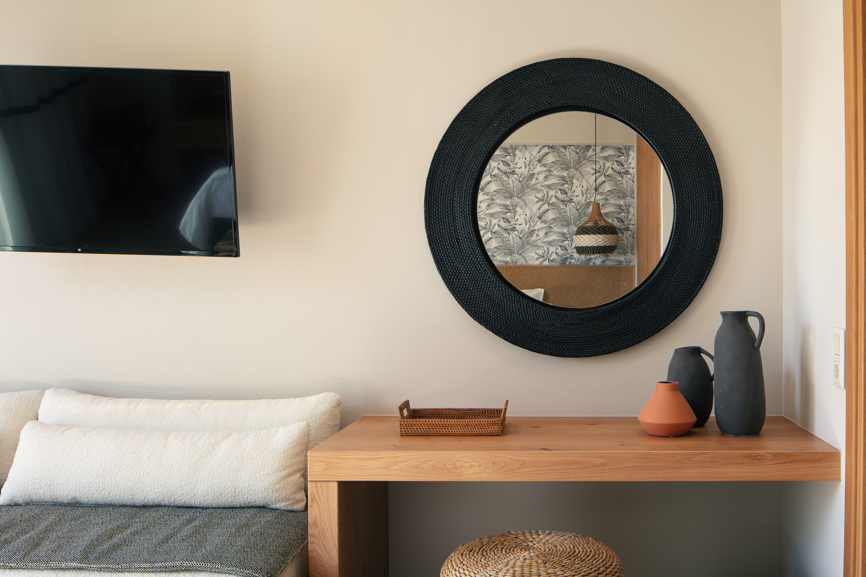 Una habitación con una televisión montada en la pared, un espejo redondo, una mesita consola de madera con objetos decorativos y una cesta tejida.