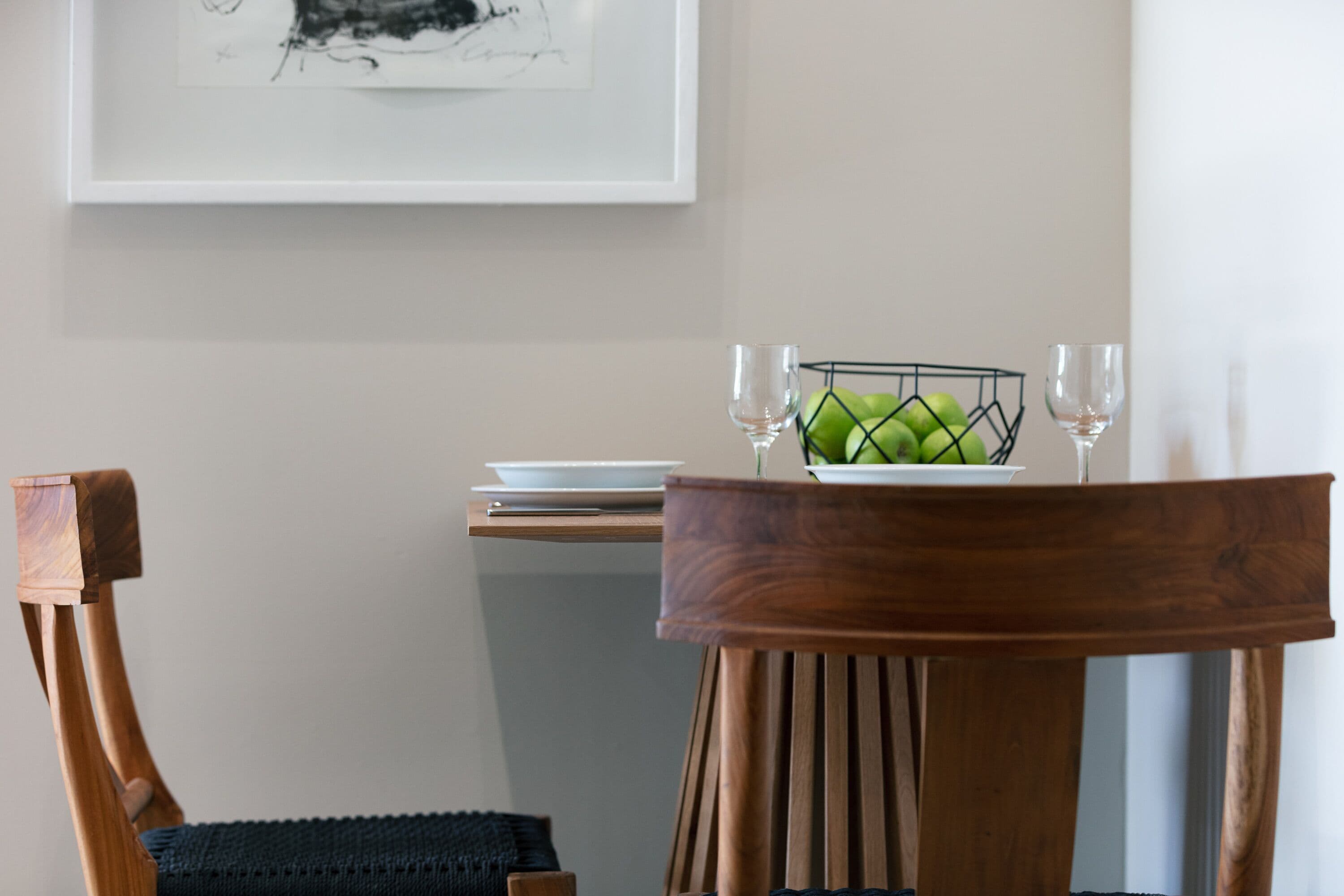 A dining area with a wooden chair, a table with a bowl of green apples, two wine glasses, and a framed artwork on the wall.