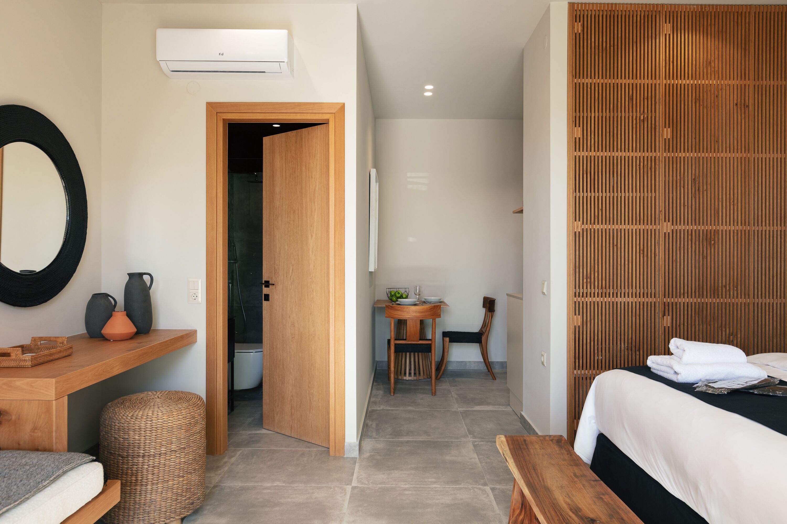 A modern hotel room with a bed, a wooden wardrobe, a small dining table with chairs, a mirror, and a wall-mounted air conditioner.