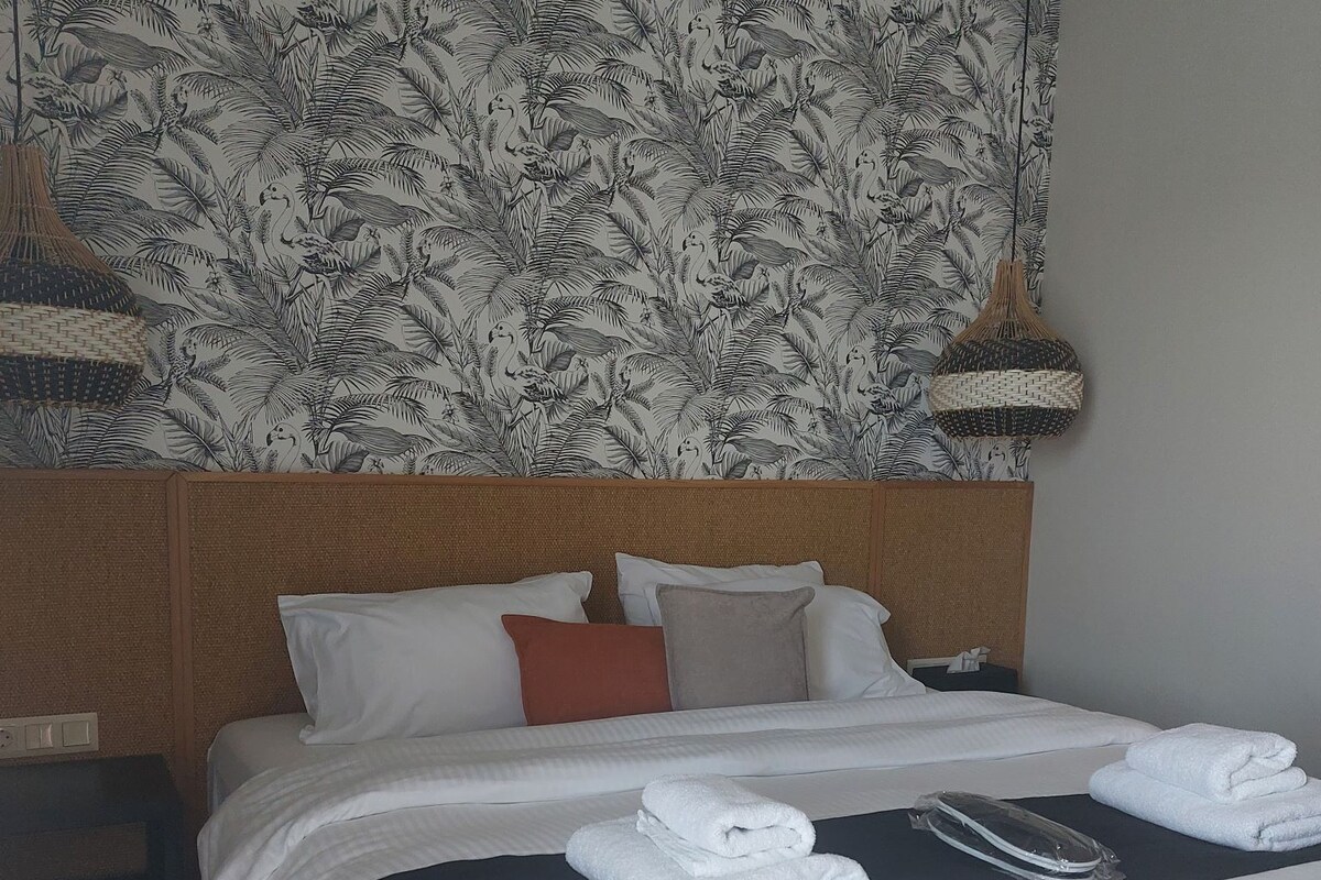 A hotel room with a large bed, a headboard with a floral pattern, two bedside lamps, and a wall with a tropical leaf design.