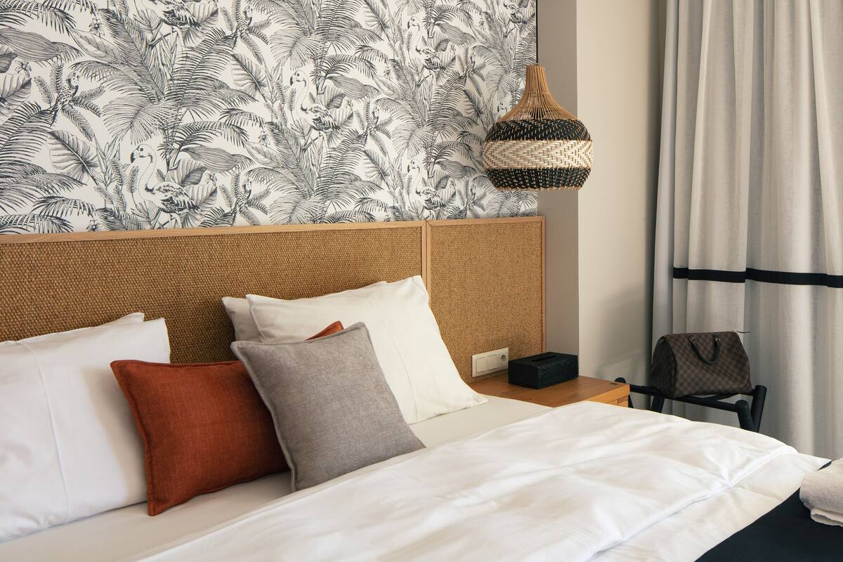 A neatly made bed with white and gray bedding, a woven headboard, and a patterned wall behind it.