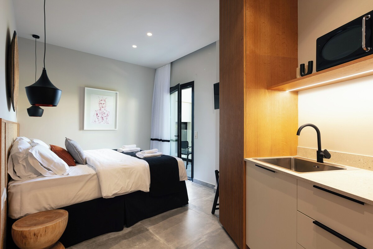 A modern hotel room with a bed, bedside table, and a built-in kitchen with a sink and microwave.