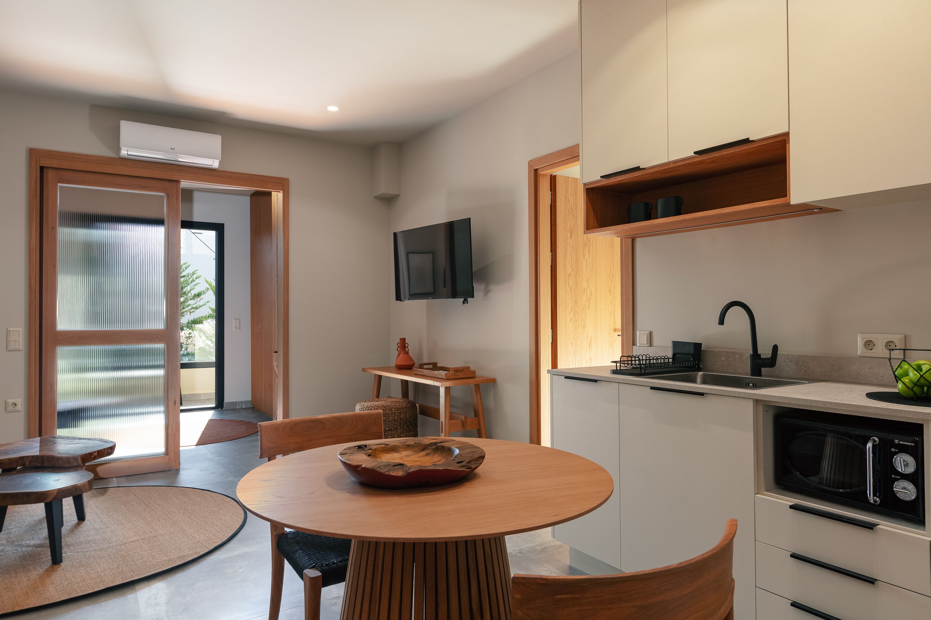 A modern kitchen with a dining area, a TV mounted above the counter, and a view through the glass door.