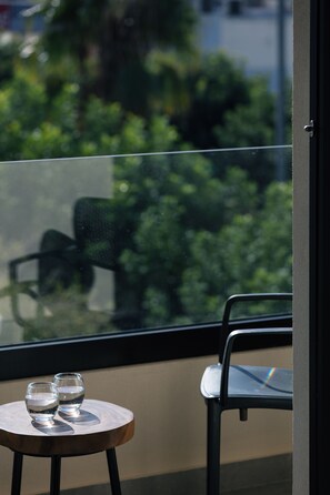 A balcony with a small round table holding two glasses of water, a chair, and a view of greenery through a glass door. - Chapter 02 Gallery Suites (Heraklion)
